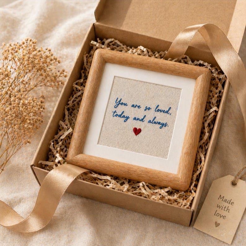 May include: A framed artwork with the text "You are so loved, today and always" and a red heart, presented in a gift box with ribbon and a tag that says "Made with love". The frame is wood.