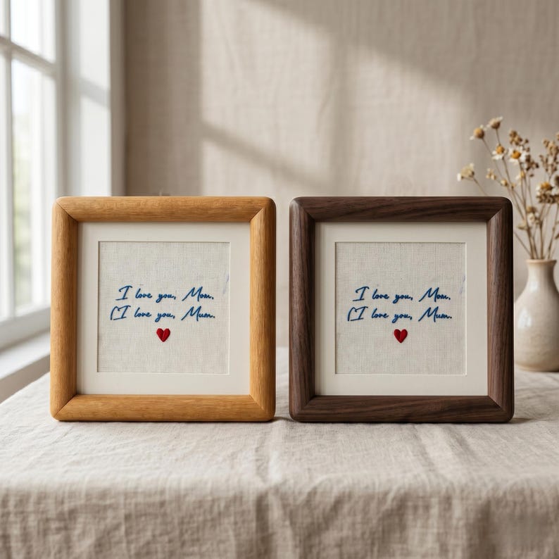 May include: Two square framed artworks with the text "I love you, Mom" and "I love you, Mum" in blue script, with a red heart. One frame is light wood, the other is dark wood. The artworks are on a neutral linen background.