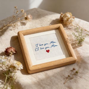 May include: A framed embroidery with the text "I love you, Mom, I love you, Mum." in blue thread. A small red heart is stitched below the text. The frame is made of light-colored wood, and the background is a neutral fabric.