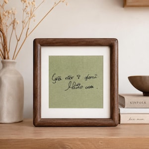 Può includere: Una cornice quadrata in legno contenente un pannello in tessuto verde salvia con testo ricamato in nero che recita "Grá mór i gcónaí Máire xxx". Il ricamo presenta uno stile di scrittura a mano con un piccolo simbolo di cuore.