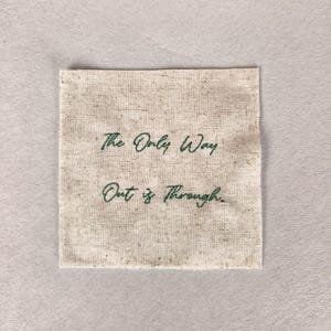 Può includere: Una toppa quadrata di tessuto beige con la frase ricamata, "The Only Way Out is Through", in corsivo verde. Il tessuto ha un aspetto strutturato ed è posto su uno sfondo neutro.