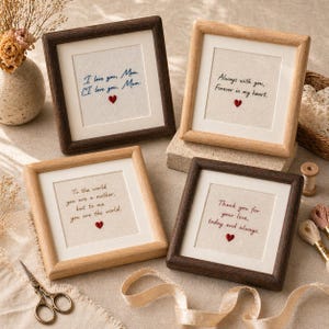May include: Four framed embroidered artworks with heartfelt messages. The frames are in various wood tones, with white matting and off-white fabric. Each piece features a red heart and text like "I love you, Mom" and "Thank you for your love."