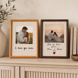 Puede incluir: Dos marcos de fotos rectangulares con paspartú de tela clara. El marco izquierdo tiene un acabado de madera clara y presenta el texto bordado 'I love you more' bajo una foto de una pareja. El marco derecho tiene un acabado de madera oscura y presenta el texto bordado 'Lots of Love from Mum & Dad' con un corazón rojo bajo una foto familiar.