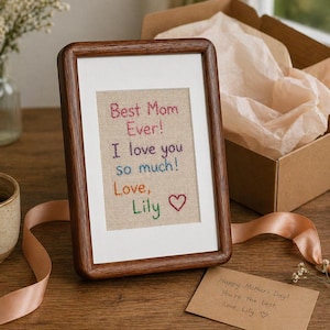 Puede incluir: Una obra de arte bordada enmarcada con el texto "Best Mom Ever! I love you so much! Love, Lily" en hilo colorido. El marco es de madera oscura, y una pequeña tarjeta con un mensaje escrito a mano es visible.