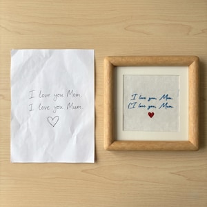 May include: A framed embroidery with the text "I love you, Mom" and "I love you, Mum" in blue thread, with a small red heart. Next to it is a handwritten note with the same text and a heart.