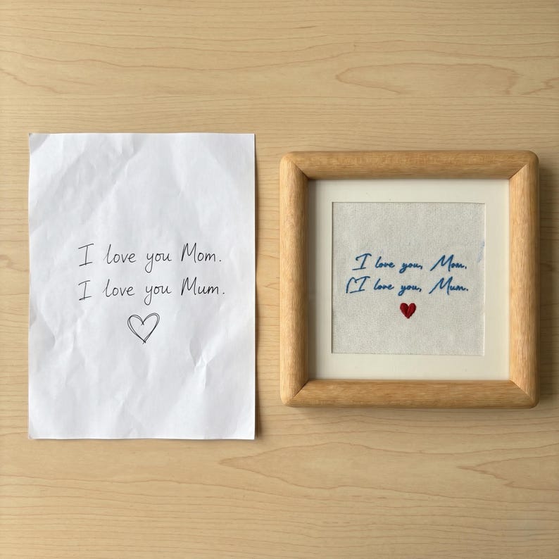 May include: A framed embroidery with the text "I love you, Mom" and "I love you, Mum" in blue thread, with a small red heart. Next to it is a handwritten note with the same text and a heart.