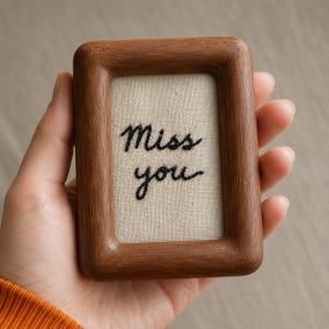 Pode incluir: Uma pequena moldura retangular de madeira com cantos arredondados. A moldura contém um tecido bege com as palavras bordadas "Miss you" em fio preto. A moldura é segurada em uma mão.