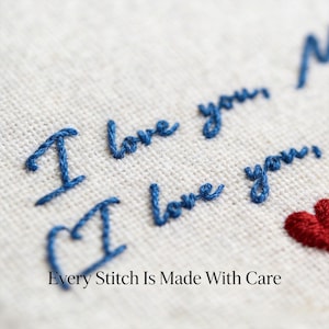 May include: Close-up of embroidered text on a light-colored fabric. The words "I love you" are stitched in blue thread, with a red heart and the phrase "Every Stitch Is Made With Care" below. The embroidery is detailed and handmade.