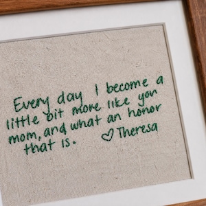May include: A framed embroidery piece on light beige fabric featuring dark green stitched text that reads: Every day I become a little bit more like you mom, and what an honour that is. A small heart symbol precedes the name Theresa.