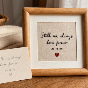May include: A framed artwork with the text "Still us, always love forever" and the date 06.12.24, along with a small embroidered red heart. A matching card with the same text and a heart is also present.