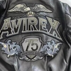 Puede incluir: Chaqueta de cuero negro con parches bordados. Presenta la palabra "AVIREX" en letras grandes, un emblema de &aacute;guila y un parche "75 AVIATOR'S KING". La chaqueta tiene un estilo bombardero cl&aacute;sico.