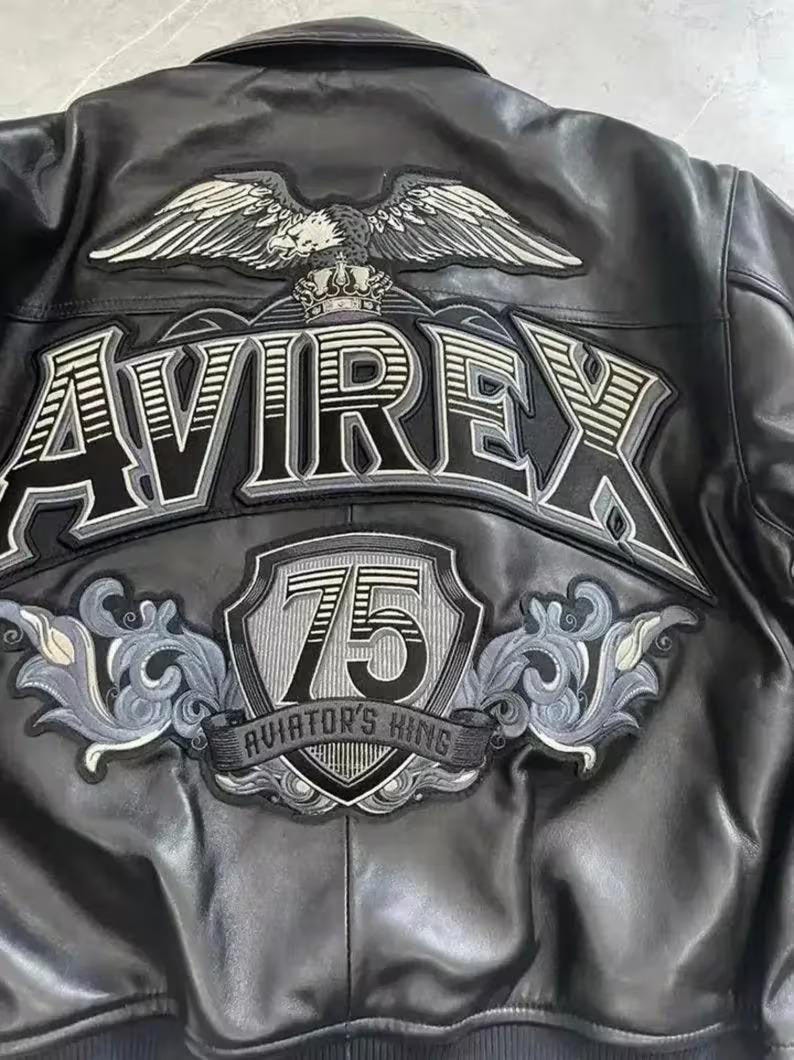 Puede incluir: Chaqueta de cuero negro con parches bordados. Presenta la palabra "AVIREX" en letras grandes, un emblema de &aacute;guila y un parche "75 AVIATOR'S KING". La chaqueta tiene un estilo bombardero cl&aacute;sico.