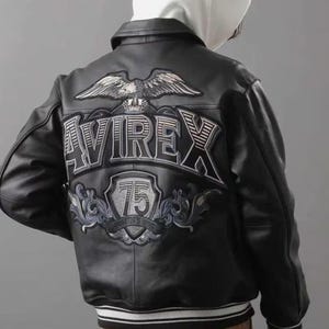 Puede incluir: Chaqueta de cuero negra con dobladillo a rayas blancas y negras. La parte trasera presenta un gran dise&ntilde;o bordado con la palabra "AVIREX", un &aacute;guila y el n&uacute;mero "75".