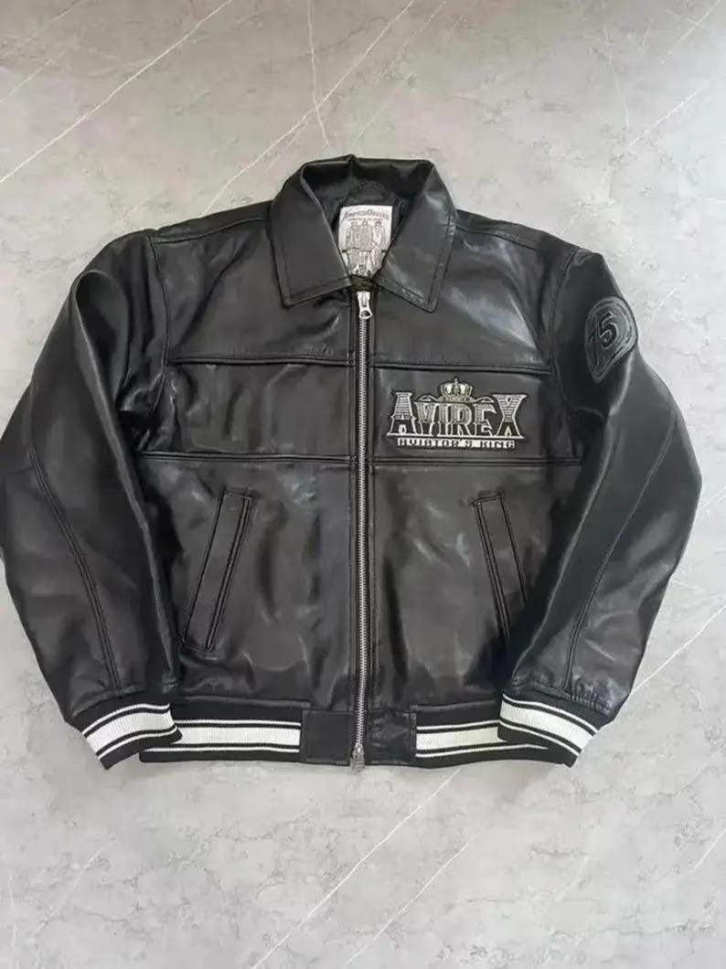 Puede incluir: Chaqueta de cuero negra con cremallera, cuello y pu&ntilde;os y dobladillo a rayas blancas. La chaqueta presenta el logotipo "AVIREX" en el pecho y un parche con el n&uacute;mero "5" en la manga. Un estilo bombardero cl&aacute;sico.