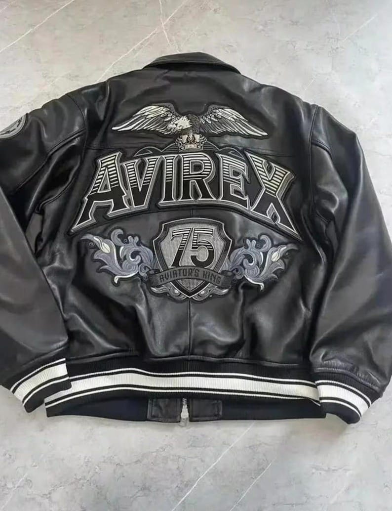 Puede incluir: Chaqueta de cuero negro con pu&ntilde;os y dobladillo a rayas blancas. La parte trasera presenta un gran dise&ntilde;o bordado con la palabra "AVIREX", un emblema de &aacute;guila y el n&uacute;mero "75". La chaqueta tiene cierre de cremallera.