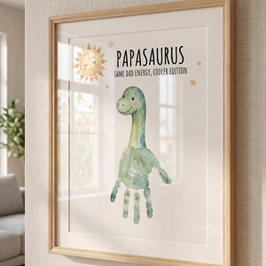 May include: Framed art print featuring a watercolor dinosaur made from a handprint. The print includes the text "Papasaurus" and "Same Dad Energy, Cooler Edition." A smiling sun and stars decorate the top of the print. The frame is light brown.