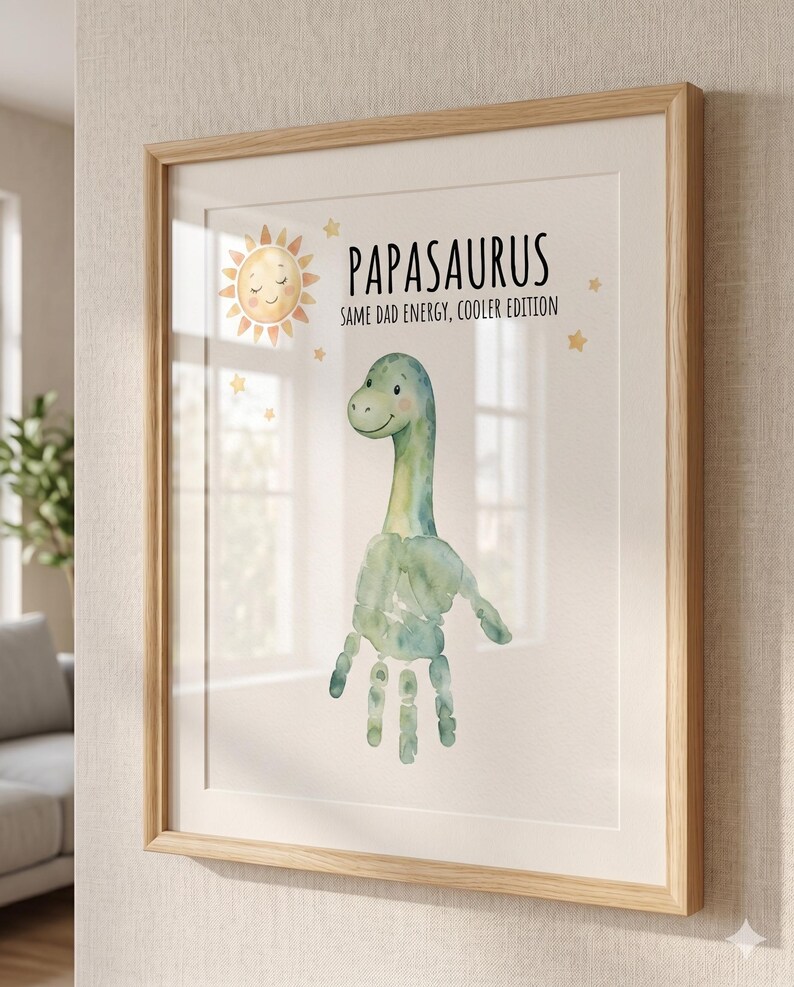 May include: Framed art print featuring a watercolor dinosaur made from a handprint. The print includes the text "Papasaurus" and "Same Dad Energy, Cooler Edition." A smiling sun and stars decorate the top of the print. The frame is light brown.