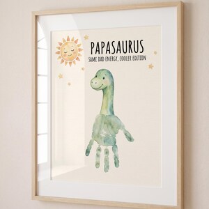 May include: Framed art print featuring a watercolor dinosaur created from a handprint. The print includes a smiling sun, stars, and the text "PAPASAURUS SAME DAD ENERGY, COOLER EDITION" in a playful font. The frame is light wood.