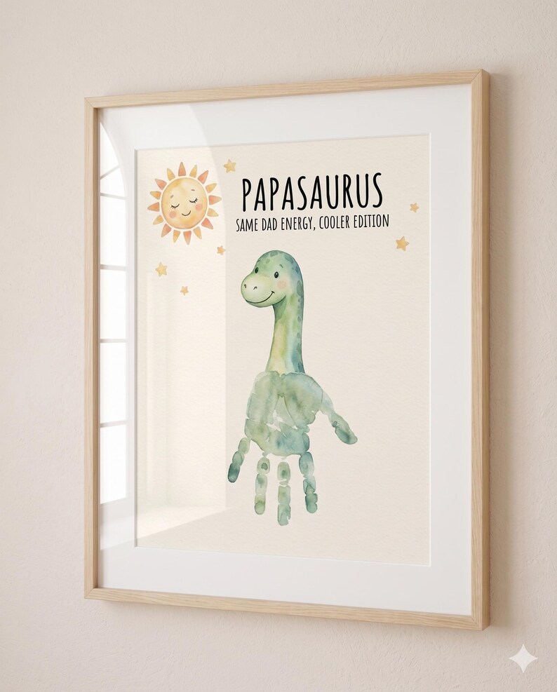 May include: Framed art print featuring a watercolor dinosaur created from a handprint. The print includes a smiling sun, stars, and the text "PAPASAURUS SAME DAD ENERGY, COOLER EDITION" in a playful font. The frame is light wood.