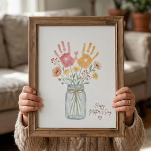 Könnte beinhalten: Ein gerahmter Aquarelldruck mit Handabdrücken als Blumen in einem Einmachglas. Das Kunstwerk enthält den Text "Happy Mother's Day" mit einem Herz, in einem Holzrahmen.