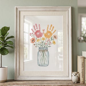 Könnte beinhalten: Gerahmtes Aquarellbild mit einem Blumenstrauß und Handabdrücken in einem Einmachglas. Die Handabdrücke sind in Rosa und Orange gehalten, mit gelben, orangefarbenen und roten Blumen. Der weiße Rahmen hat eine Used-Optik.