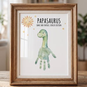 May include: A framed print with a watercolor dinosaur design. The print features a green dinosaur with a handprint body and the text "Papasaurus: Same Dad Energy, Cooler Edition." The frame is made of wood.