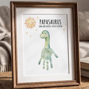 May include: Framed art print featuring a watercolor dinosaur made from a handprint. The print includes the text "Papasaurus: Same Dad Energy, Cooler Edition" with a smiling sun and stars. The frame is a warm brown color.