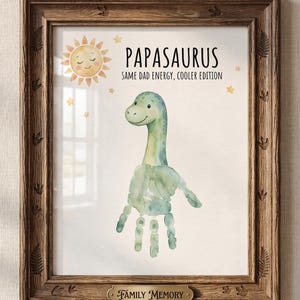 May include: A framed print featuring a watercolor dinosaur made from handprints, with the text "Papasaurus: Same Dad Energy, Cooler Edition." The print includes a smiling sun and stars, with a "Family Memory" plaque.