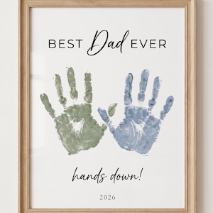 May include: Framed print with the text "BEST Dad EVER" above two handprints, one green and one blue. Below the handprints is the phrase "hands down!" and the year "2026". The frame is a light brown color.