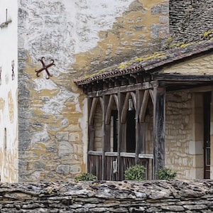 May include: Exterior of a stone building with a wooden porch and a tiled roof. The building has a weathered appearance with a mix of white and yellow tones. A decorative iron cross is mounted on the wall.