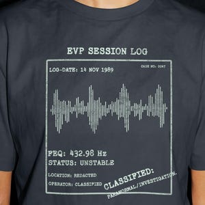 May include: A dark grey short-sleeve t-shirt featuring a white graphic print of an EVP SESSION LOG. The design includes a sound wave, log-date 14 NOV 1989, case no 0067, feq 432.98 Hz, status unstable, location redacted, and classified paranormal investigation.