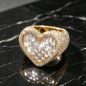 May include: A gold-toned heart-shaped ring featuring a center filled with baguette-cut clear stones and a border encrusted with small round clear stones. The entire surface of the ring band is also covered in tiny round clear stones.