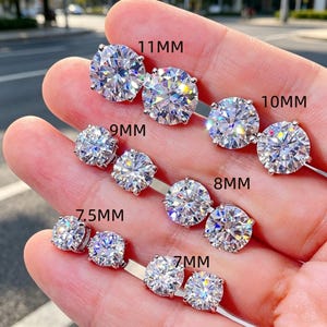 May include: A collection of round, brilliant-cut clear stud earrings arranged in pairs on a hand. The pairs are labeled with sizes 11mm, 10mm, 9mm, 8mm, 7.5mm, and 7mm. Each earring features a multi-faceted stone set in a silver-toned metal prong setting.