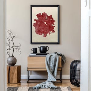 May include: A framed abstract art print featuring a red, organic design. The artwork is above a wooden cabinet with a black teapot, cup, and a grey blanket. A decorative vase with branches and a black wicker stool are also present.