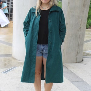 May include: A woman wearing a teal long-sleeved jacket over a black shirt and denim shorts. She is standing on a concrete surface.