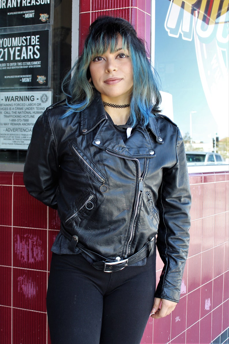 May include: A woman in a black leather biker jacket with silver zips and a belt, paired with black leggings and a choker. Her dark hair has blue streaks. The background features a red tiled wall and a window with text.