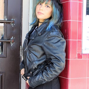 May include: A black leather jacket with a zip fastening and pockets. The jacket is worn with black trousers. The person is leaning against a red tiled wall next to a dark brown door.