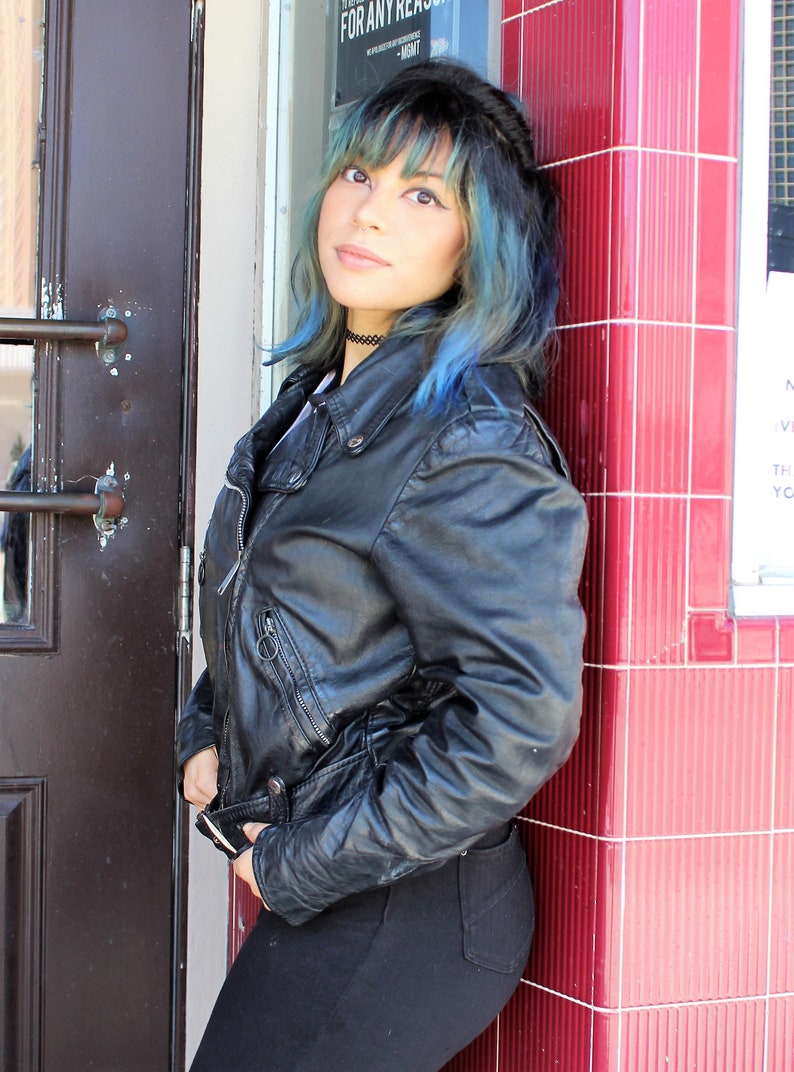 May include: A black leather jacket with a zip fastening and pockets. The jacket is worn with black trousers. The person is leaning against a red tiled wall next to a dark brown door.