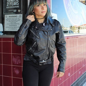 May include: Black leather biker jacket with silver zips and a belt. The jacket features a classic design with a notched collar and shoulder epaulettes. The image also shows black leggings and a black choker.