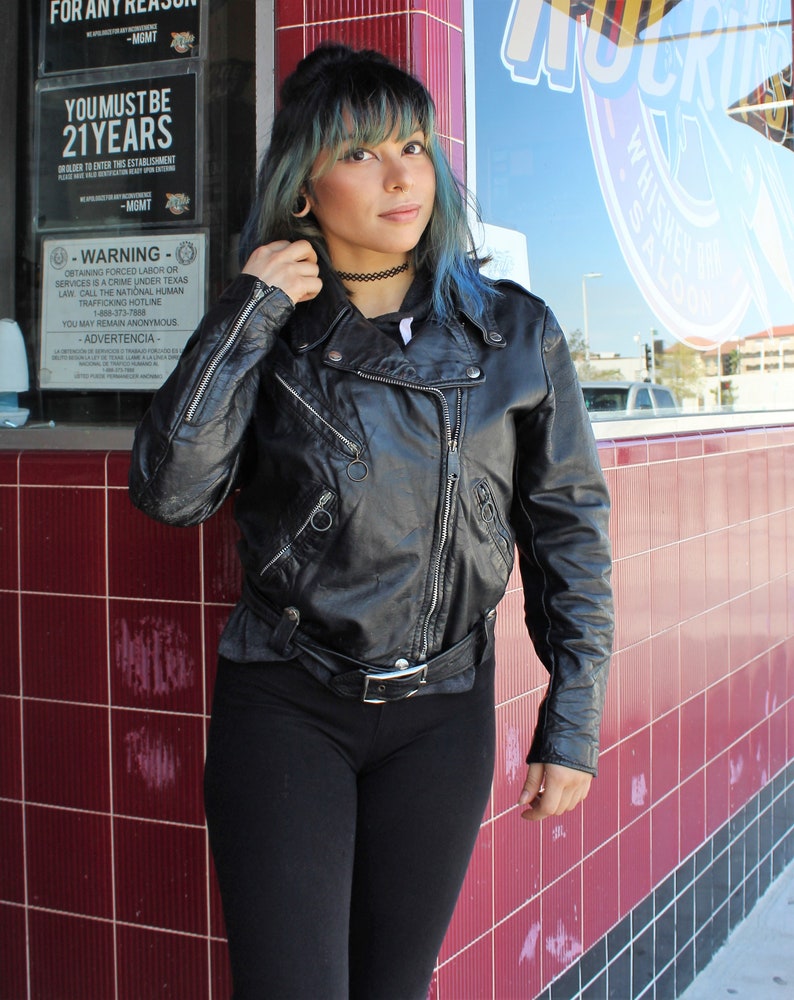 May include: Black leather biker jacket with silver zips and a belt. The jacket features a classic design with a notched collar and shoulder epaulettes. The image also shows black leggings and a black choker.
