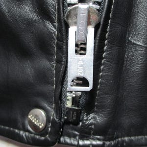 May include: Close-up of a black leather jacket featuring a silver zipper and a snap closure. The zipper pull is branded with "ZORR". The leather has a textured appearance, suggesting a vintage style.