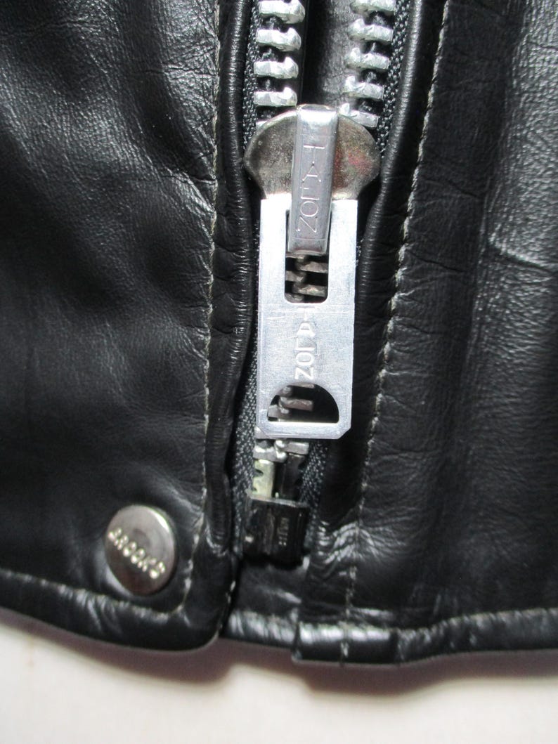 May include: Close-up of a black leather jacket featuring a silver zipper and a snap closure. The zipper pull is branded with "ZORR". The leather has a textured appearance, suggesting a vintage style.