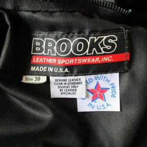 May include: A close-up of a black garment featuring a label that reads "BROOKS LEATHER SPORTSWEAR, INC. MADE IN U.S.A." The label also indicates a size 30. Another label states "GENUINE LEATHER CLEAN IN STODDARD SOLVENT ONLY BY LEATHER SPECIALIST".