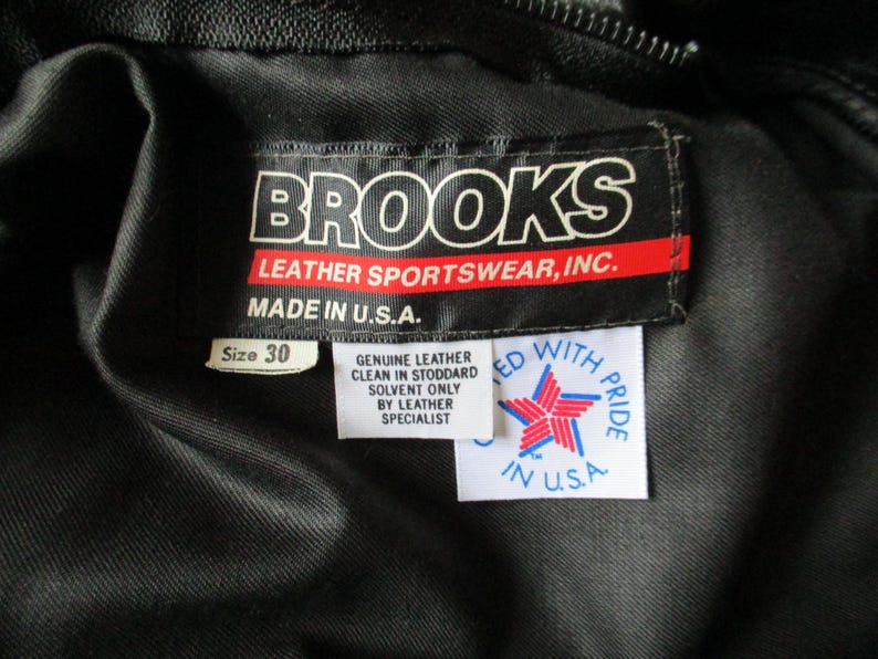 May include: A close-up of a black garment featuring a label that reads "BROOKS LEATHER SPORTSWEAR, INC. MADE IN U.S.A." The label also indicates a size 30. Another label states "GENUINE LEATHER CLEAN IN STODDARD SOLVENT ONLY BY LEATHER SPECIALIST".