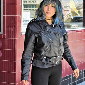 May include: Black leather biker jacket with silver zips and a belt. The jacket is worn over a grey top and paired with black leggings. The background features a red tiled wall and a storefront.