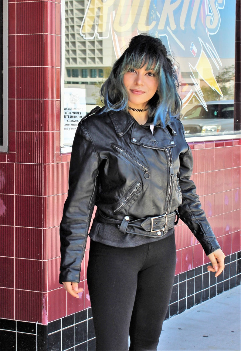 May include: Black leather biker jacket with silver zips and a belt. The jacket is worn over a grey top and paired with black leggings. The background features a red tiled wall and a storefront.