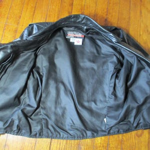 May include: Black leather jacket with a zip fastening, open to show a black satin lining. The jacket has two front pockets and a label that reads "BROOKS". The jacket is laid flat on a wooden surface.