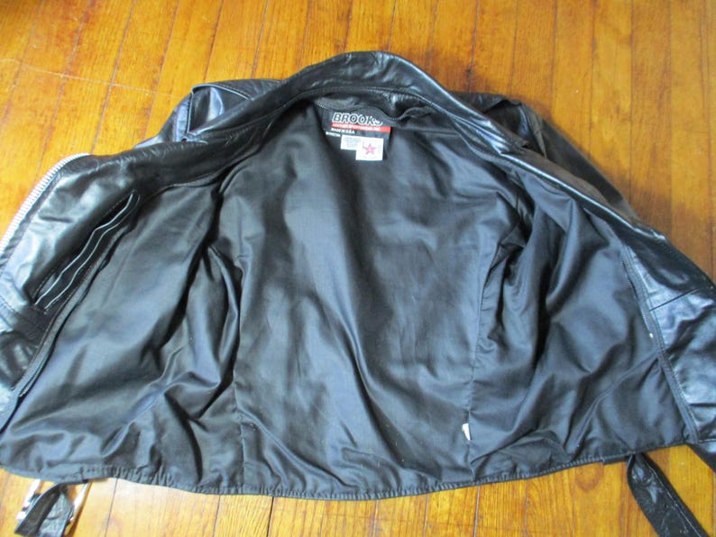May include: Black leather jacket with a zip fastening, open to show a black satin lining. The jacket has two front pockets and a label that reads "BROOKS". The jacket is laid flat on a wooden surface.