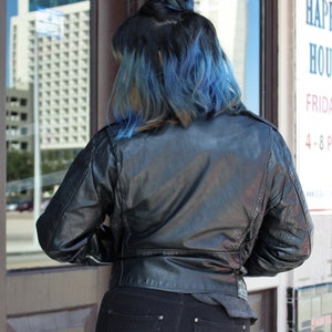 May include: A black leather jacket with a classic design. The jacket has a zip closure and a tailored fit. The person is wearing black jeans. The person's hair is dark with blue highlights. The image is taken outdoors.