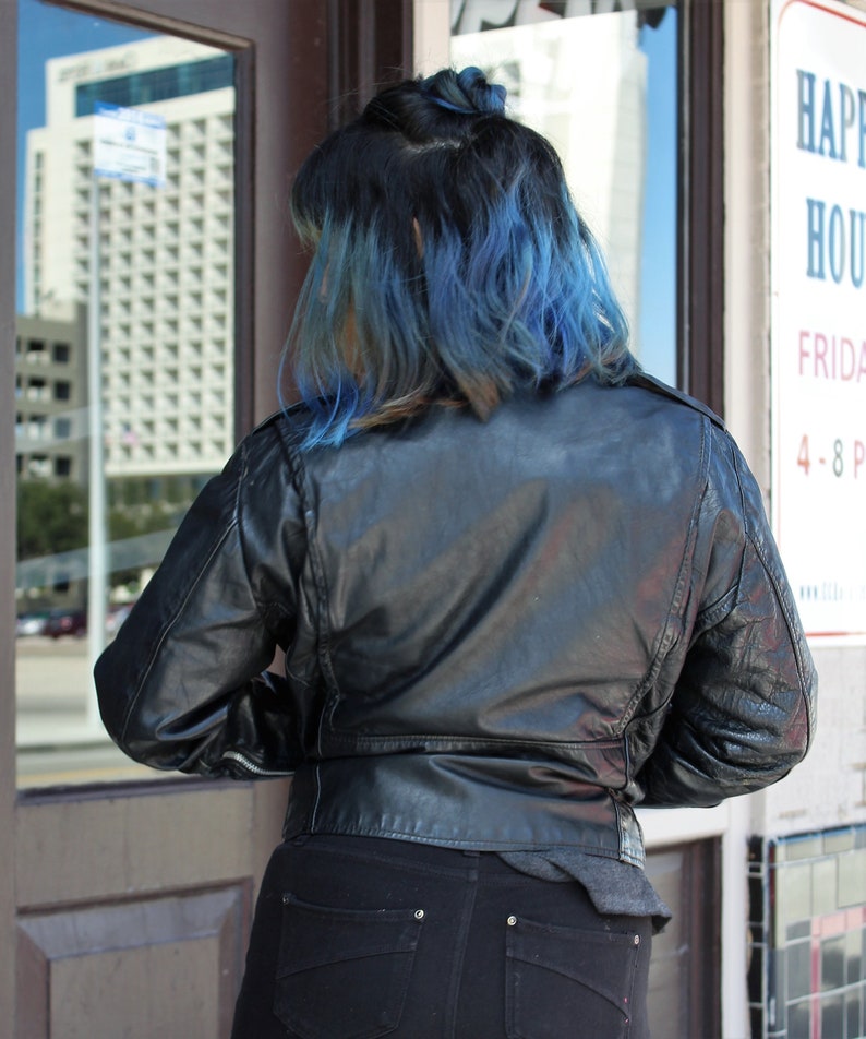 May include: A black leather jacket with a classic design. The jacket has a zip closure and a tailored fit. The person is wearing black jeans. The person's hair is dark with blue highlights. The image is taken outdoors.
